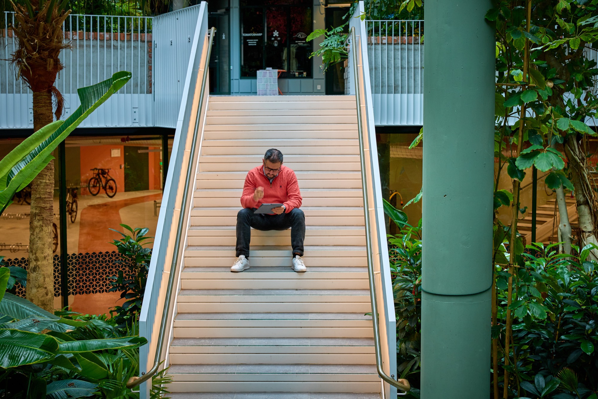 A product manager sits partway up a wide staircase in a plant-filled office atrium, reading from a tablet.